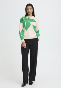 Világos rózsaszín pulóver zöld levélmintával, fekete bő szárú nadrággal párosítva. Puha anyagú, kerek nyakkivágású, bordázott mandzsettákkal.