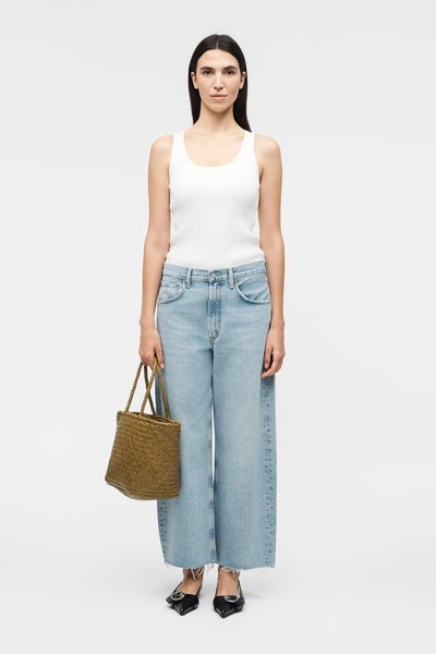 Γυναίκα στέκεται φορώντας λευκό αμάνικο τοπ, φαρδιά τζιν παντελόνια ανοιχτό μπλε χρώμα, μαύρα μυτερά παπούτσια, και κρατώντας καφέ πλεκτή τσάντα tote.
