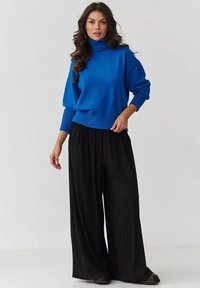 Niebieski sweter w golf z luźnym krojem i ściągaczami, w połączeniu z szerokimi czarnymi spodniami i płaskimi butami. Gładka tekstura, casualowy design.