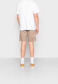 Shorts beiges avec une coupe décontractée et un design uni, associés à un t-shirt blanc et des chaussettes de couleur claire, vus de derrière.