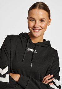 Czarna bluza z kapturem z logo, z białymi akcentami w kształcie chevronu na rękawach. Wykonana z miękkiego, teksturowanego materiału.