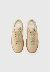 Sneaker in suede beige con suole in gomma marrone chiaro. Caratteristiche includono un design a punta tonda, lacci piatti e un marchio discreto sul lato.