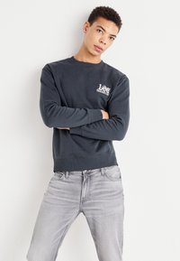 Sudadera gris oscuro de algodón, con cuello redondo, puños de canalé y un logo blanco en el lado izquierdo del pecho. Combinada con unos vaqueros gris claro.
