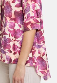 Kwiatowa bluzka w odcieniach różu i fioletu, charakteryzująca się miękką fakturą, krótkimi rękawami i zaokrąglonym dołem. Wzór zawiera duże kwiaty i liście.