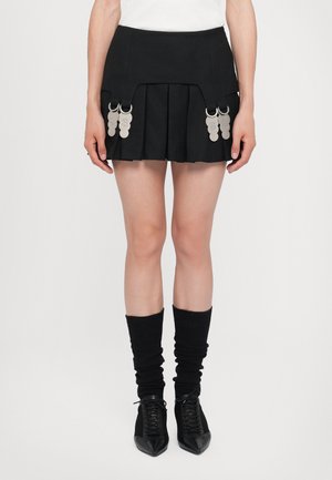 Jupe plissée noire avec des anneaux métalliques attachés. Associée à des chaussettes noires montantes et des chaussures noires pointues avec une finition texturée.