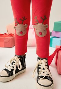 Pantaloni da jogging rossi con applicazioni a forma di faccia di renna sulle ginocchia, abbinati a sneakers alte nere con lacci bianchi e suole in gomma.