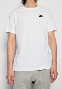 White cotton t-shirt with short sleeves, round neckline, and a small black Nike logo on the left chest area. Smooth texture.