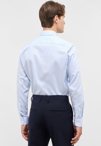 Uomo che indossa una camicia a maniche lunghe a strisce blu chiaro e bianche infilata in pantaloni tailleur blu scuro, di spalle su sfondo grigio uniforme.