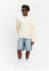 Lichtbeige gebreide trui met een losse pasvorm, gecombineerd met lichtblauwe cargoshorts met meerdere zakken. Witte sneakers en enkelsokken zichtbaar.