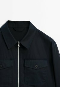 Veste noire avec col, fermeture éclair à l'avant et deux poches poitrine à rabat boutonné, présentée sur fond blanc.