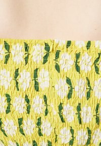 Tecido amarelo com pregas, apresentando um padrão floral com flores brancas e folhas verdes. Textura suave com detalhes franzidos e elásticos.