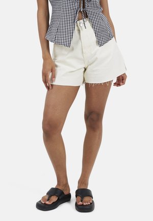 Kobieta w białych szortach jeansowych, czarno-białym kraciastym krótkim topie i czarnych sandałach na platformie, stojąca na białym tle.