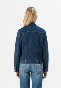 Blond person som vänder ryggen till, iklädd en mörkblå denimjacka och ljusblå jeans med sydda bakfickor mot en enkel vit bakgrund.