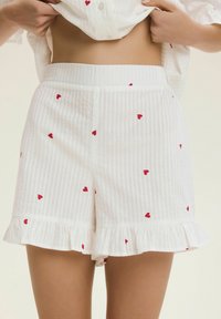 Białe spodenki w paski z czerwonymi wzorami serc, z falbaniastym brzegiem i elastycznym paskiem w talii dla wygody. Wykonane z lekkiej tkaniny.
