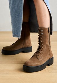 Bottines en suédé marron avec une semelle épaisse noire, dotées d'un laçage à l'avant et d'une fermeture éclair sur le côté, posées sur un sol en bois.