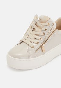Beige lakota ādas sporta apavi ar apaļu degumu, slāņainiem šņorēm, zelta toņa rāvējslēdzēja akcentiem un faktūru balto zoli. Redzama mīksta oderīte.