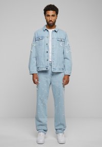 Light blue denim jacket with distressed details, paired with matching light blue jeans featuring paint splatter accents and white sneakers.