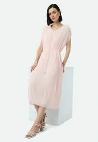 Helles rosa plissiertes Kleid mit V-Ausschnitt und kurzen Ärmeln, tailliert mit einem Band. Kombiniert mit weißen Riemchensandalen.