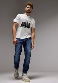 Valkoinen graafinen t-paita, jossa on musta kaupunkisilhuetti-printti, yhdistettynä sinisiin farkkuihin ja beigeklassisiin tennareihin, joissa on teksturoidut paneelit ja nauhat.