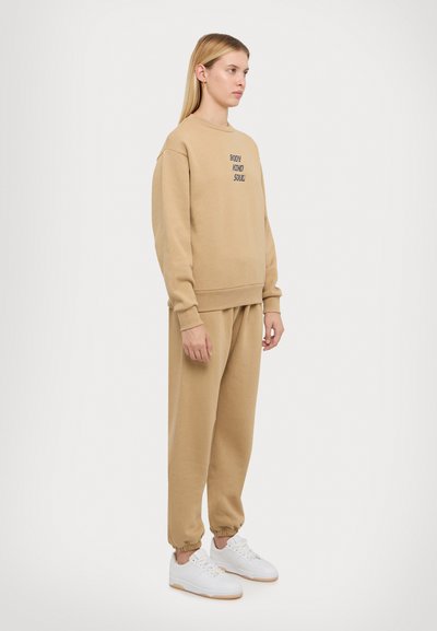Beige sweatshirt en bijpassende joggers, katoenmix, relaxte pasvorm. Sweatshirt heeft geborduurde tekst; joggers hebben elastische boorden. Witte sneakers.