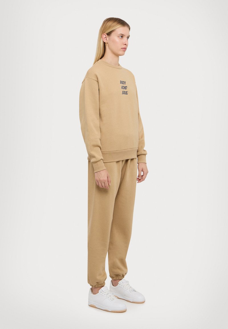 Beige sweatshirt and matching joggers, cotton blend, relaxed fit. Sweatshirt has embroidered text; joggers have elastic cuffs. White sneakers.