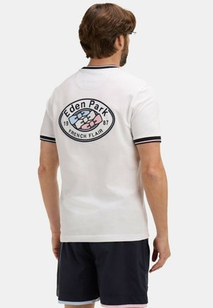 Homme portant un polo blanc avec le logo "Eden Park French Flair" et un motif de nœud papillon dans le dos, associé à un short noir.