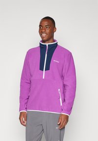 Fleece trøje i livlig lilla med en kontrasterende marineblå kvart-zip krave og to sidelommer med lynlås. Har hvide detaljer og logo.
