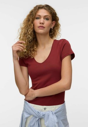 Kvinde med krøllet blondt hår iført en tætsiddende vinrød V-hals t-shirt og en lyseblå skjorte bundet om taljen, poserer mod en ensfarvet baggrund.