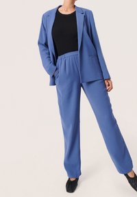 Blå skräddarsydd kavaj med en enda knapp, ihop med avslappnade byxor med elastisk midja. Toppen är en figurnära svart ribbad stickad tröja. Slät textur.