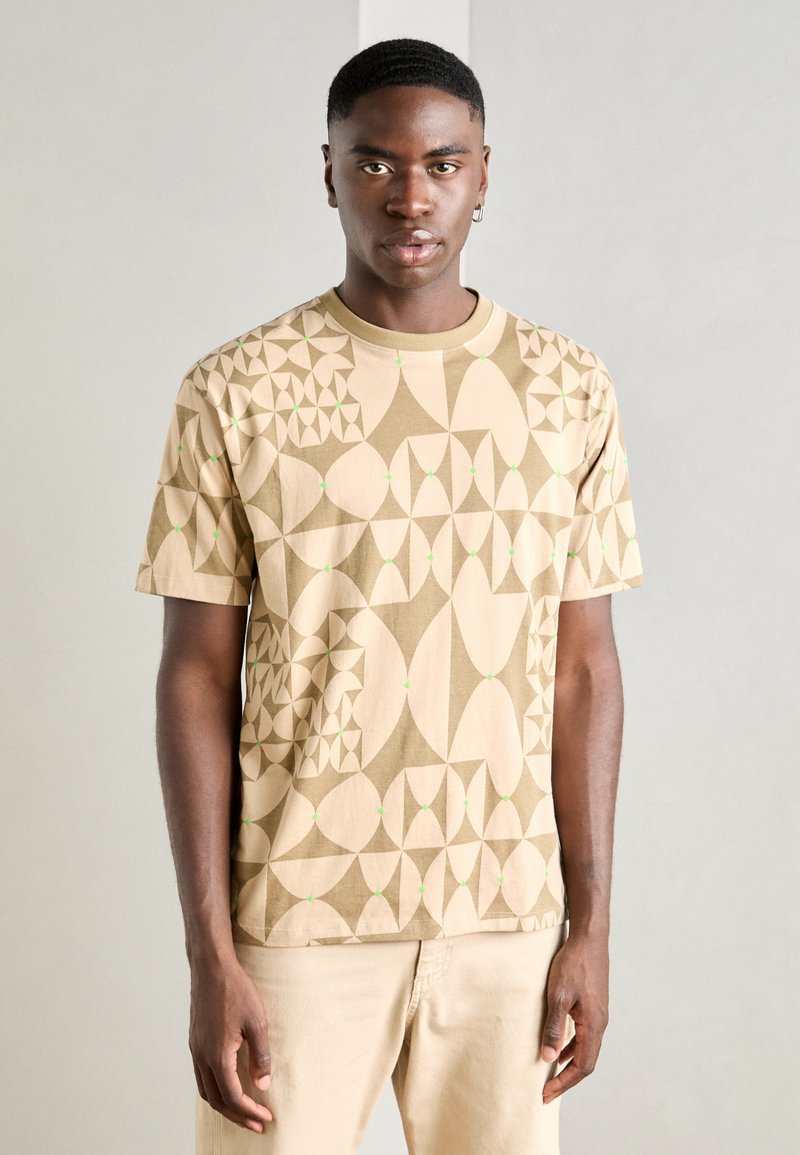 Beige Baumwoll-T-Shirt mit geometrischen Mustern in hellem und dunklem Beige sowie kleinen grünen Punkten. Mit kurzen Ärmeln und einem Rundhalsausschnitt.
