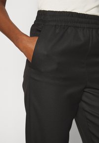 Main à la peau brune reposant dans la poche d’un pantalon noir à taille élastique porté avec un haut blanc.