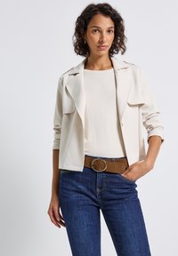 Femme aux cheveux bouclés, portant un blazer court beige, un haut beige clair, un jean bleu et une ceinture en daim marron avec une grande boucle ronde.