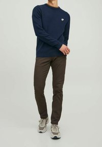 Dunkelblaues Sweatshirt mit Rundhalsausschnitt und kleinem Logo, kombiniert mit dunkelbraunen Hosen und grauen Turnschuhen mit weißen Akzenten. Sichtbare Nähte.