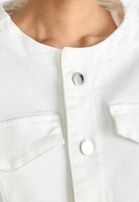 Valkoinen farkkutakki, jossa on pyöreä pääntie, hopeiset napit ja kaksi rintataskua läpällä. Suora pinta ja rento muotoilu.