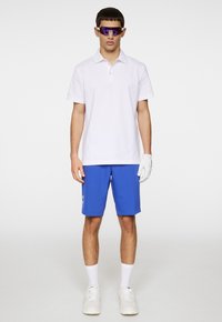 Vit pikétröja, blå kortbyxor, vita sneakers, vita ankelsockor och lila solglasögon. Kläderna kompletteras med en vit handske.