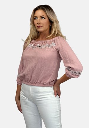 Helles pinkes Langarmbluse mit elastischem Saum, verziert mit weißer Spitze am Ausschnitt und an den Bündchen. Getragen mit weißen Hosen.