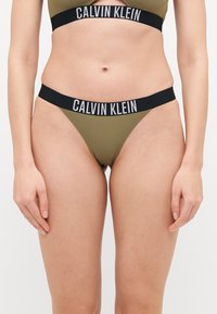 Ελαιόλαδο πράσινο μπικίνι Calvin Klein με μαύρες λογότυπες ελαστικές ζώνες. Το τοπ έχει σχέδιο με κοψίματα και το κάτω μέρος είναι χαμηλής μέσης στυλ στρινγκ.