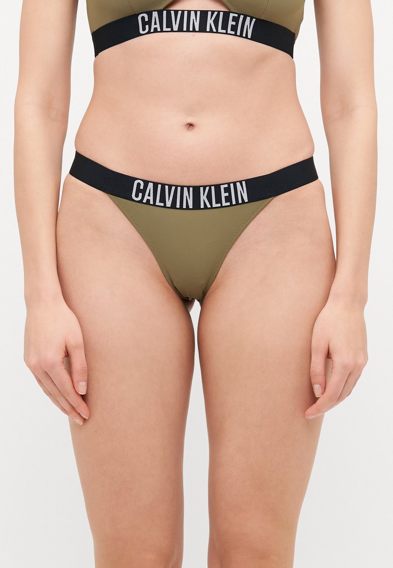 Ελαιόλαδο πράσινο μπικίνι Calvin Klein με μαύρες λογότυπες ελαστικές ζώνες. Το τοπ έχει σχέδιο με κοψίματα και το κάτω μέρος είναι χαμηλής μέσης στυλ στρινγκ.