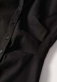 Chemise noire à manches longues avec des boutons visibles sur le devant et des plis subtils près des épaules et des manches.