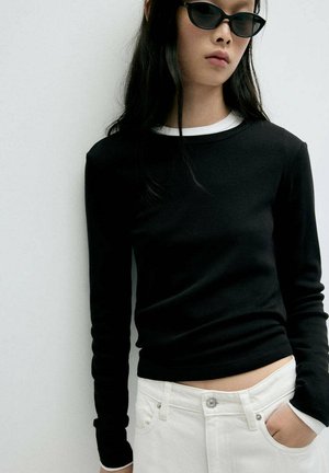 Jeune personne portant un haut noir à manches longues par-dessus une chemise blanche, un pantalon blanc et des lunettes de soleil noires, debout contre un mur clair uni.