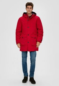 Uomo con una giacca invernale rossa con cappuccio, bottoni e tasche, jeans blu e scarpe nere, in piedi contro uno sfondo semplice.