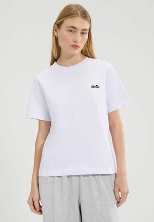 Młoda kobieta o długich blond włosach, ubrana w białą koszulkę ellesse i jasnoszare spodnie dresowe, stojąca na jednolitym tle.