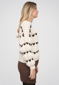 Blondýna v profilu, oblečená v béžovém pleteném svetru s černými vzory ve tvaru srdce a hnědé sukni na jednobarevném bílém pozadí.