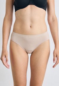Model stoji s povjerenjem, noseći crni grudnjak i bež Calvin Klein donje rublje, prikazujući minimalistički stil na neutralnoj pozadini.