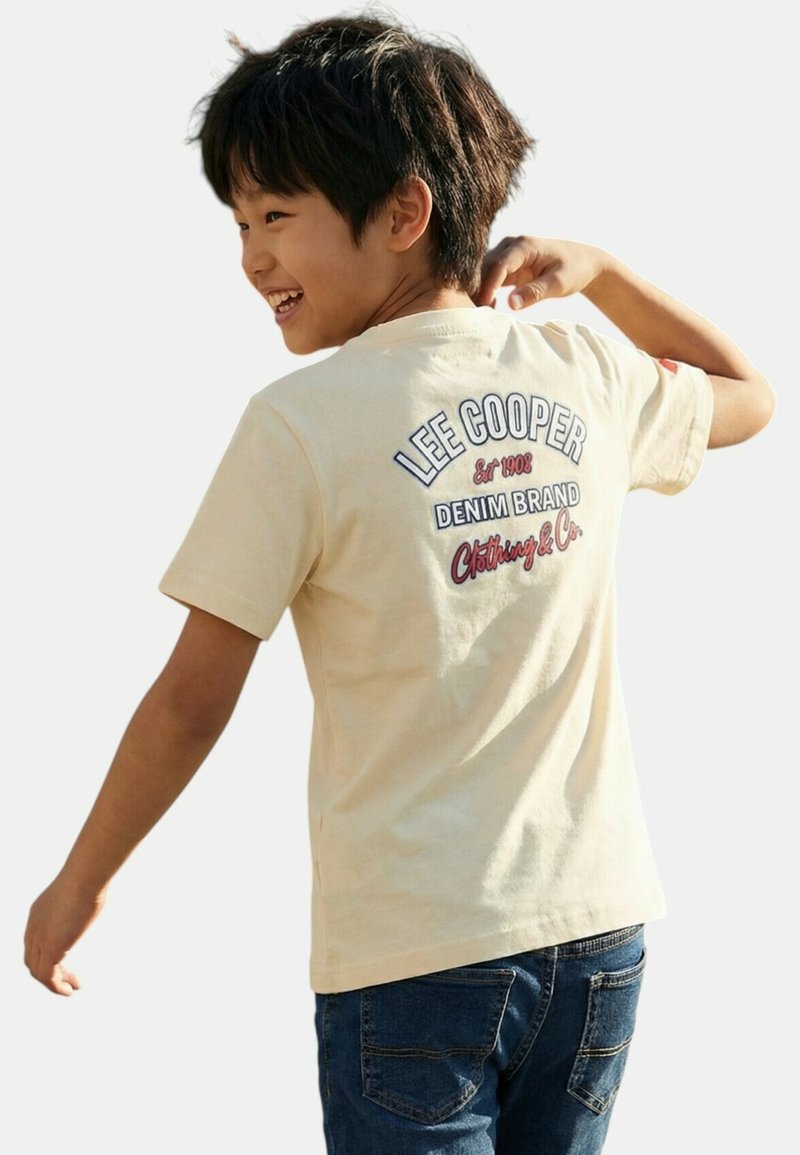 Garçon souriant se retournant, portant un t-shirt beige clair Lee Cooper avec un jean en denim, montrant le logo de la marque au dos du t-shirt.