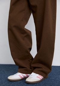 Pantalon marron à jambes larges associé à des baskets blanches avec des rayures roses. Le tissu semble lisse, de couleur unie et offre une coupe décontractée.