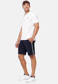 Hvit polo skjorte med korte ermer og en liten logo, kombinert med marineblå shorts med hvite striper på siden og oppbrettede ermer, brukt med hvite sneakers.