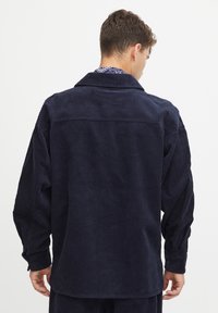 Navy corduroy shirt with a classic collar, large back panel, and drop shoulders. Features ribbed texture and buttoned cuffs.