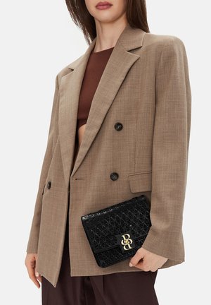 Frau in einem braunen, doppelt geknöpften Blazer, die eine strukturierte schwarze Clutch mit goldener Verschlussklammer hält und darunter ein dunkelbraunes Oberteil trägt.