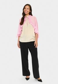 Lightweight pink cape with ruffles over a cream blouse, paired with black wide-leg trousers and pointed black shoes. Casual outfit.